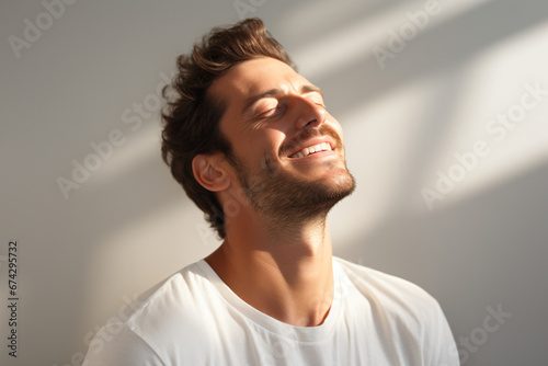Young man smiling with eyes closed and head tilted up. Breathing deeply and feeling the light that touches his face. Chiaroscuro lighting. Expression of pleasant calm on his face. On white background