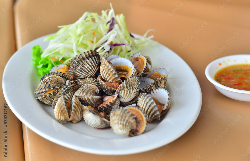 The boiled cockle with spicy sauce. This menu is set on the white plate, there are cockle, some vegetables. And it usually served with a cup of spicy, sweet and sour sauce. The boiled cockle with spicy sauce. This menu is set on the white plate, there are cockle, some vegetables. And it usually served with a cup of spicy, sweet and sour sauce.