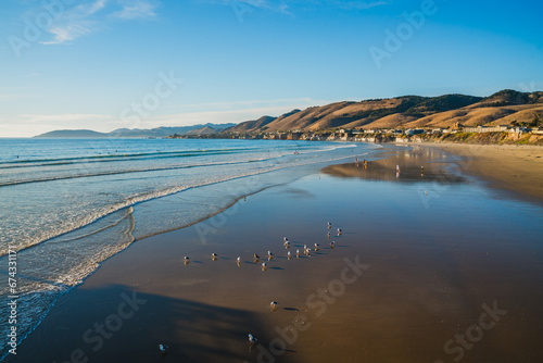 Pismo beach hills with cliffs, wide sandy beach at a low tide, dark blue ocean, and a silhouette of a town in the background at sunset, California Central Coast