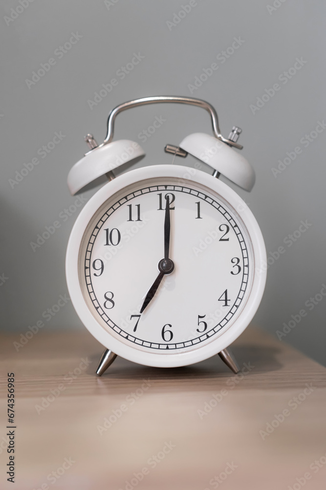 Close-up of a round white alarm clock on a table in the bedroom. The hands on the clock show seven o'clock in the morning, time to get up. Retro alarm clock on the table, vintage tone. space for text Close-up of a round white alarm clock on a table in the bedroom. The hands on the clock show seven o'clock in the morning, time to get up. Retro alarm clock on the table, vintage tone. space for text