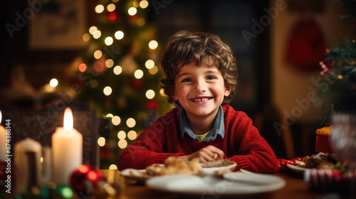 Cheerful boy in a red sweater is smiling at the Christmas dinner table, with lit candles and a decorated tree in the background, contributing to a festive atmosphere.