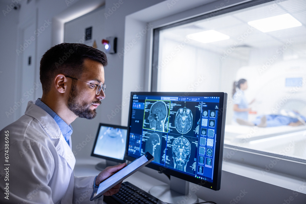 Doctor examining X-ray images on display in MRI control room while in background nurse preparing the patient for examination test. - obrazy, fototapety, plakaty 