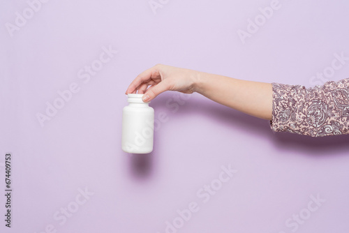 White plastic bottle in female hand in lilac background. Packaging for pills, capsules or supplements.