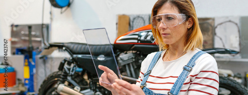Concentrated female mechanic with security glasses holding transparent tablet in front of motorcycle on garage