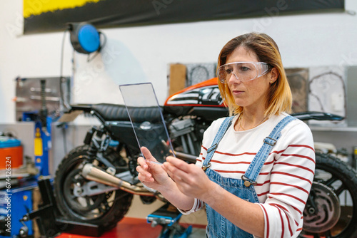 Concentrated female mechanic with security glasses holding transparent tablet in front of motorcycle on garage