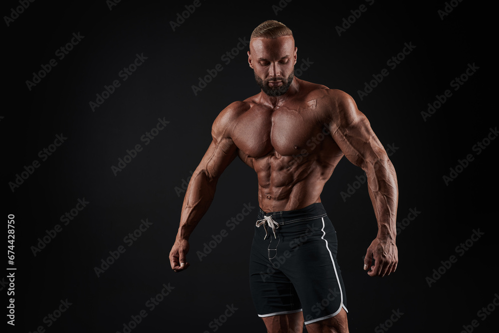 Muscular man shows his muscles against the background of a black wall. Bodybuilder, male naked torso, abs.