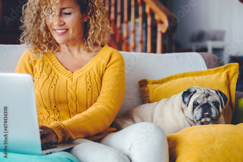 One happy woman smiling and writing on laptop with her best friend dog laying on her side both on the sofa at home. Puppy owner searching on web with computer. Indoor leisure activity together people