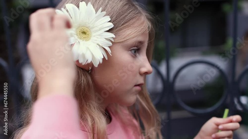 Portrait of a funny little girl emotionally talking to camera and holding a big white gerbera flower in her hair, little girl with big blue eyes funny walks on a street
