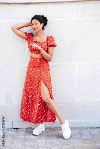 Young beautiful smiling hipster woman in trendy summer red dress. Carefree woman posing in the street near wall. Positive model outdoors in at sunny day. Cheerful and happy