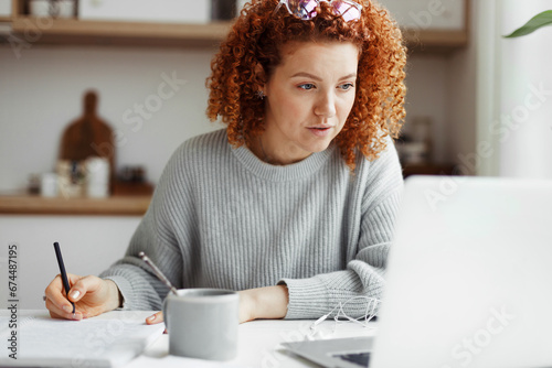 Portrait of attractive redhead caucasian businesswoman in casual clothes working from home, making business plan for launching new project, checking e-mail and financial report from her accountant