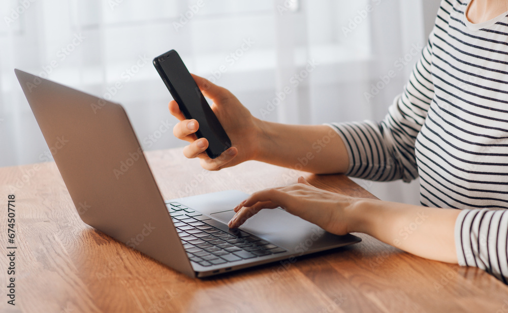 Naklejka premium Searching for information on the Internet on the phone. A girl works with a smartphone and a laptop on a table