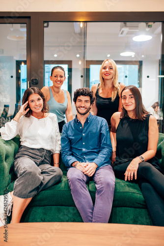 Group of optimistic young coworkers in casual outfits gathering together on comfortable sofa and looking at camera while having meeting in contemporary workspace.
