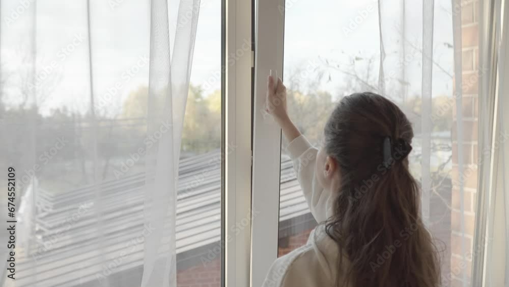 The woman parted the transparent tulle curtains and turned the handle on the PVC plastic window frame to slightly open the window and ventilate the room through a small crack. The woman parted the transparent tulle curtains and turned the handle on the PVC plastic window frame to slightly open the window and ventilate the room through a small crack.