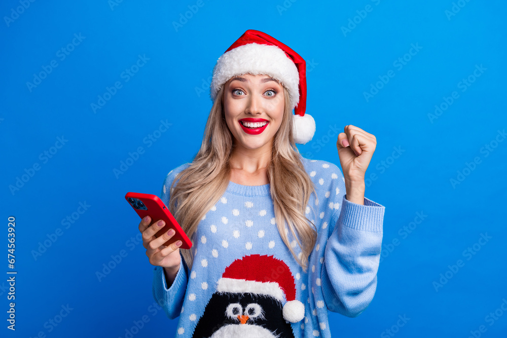 Obraz premium Photo portrait of pretty blonde young girl gadget raise fist winning dressed penguin sweater x-mas hat isolated on blue color background