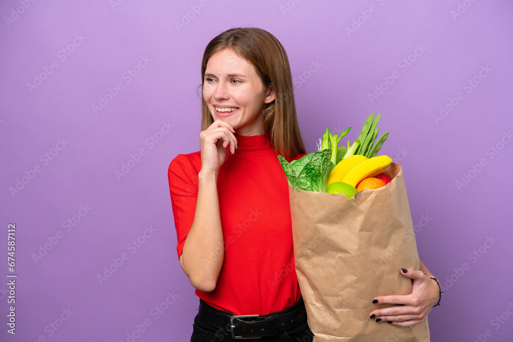 Young English woman holding a grocery shopping bag isolated on purple background looking to the side and smiling Young English woman holding a grocery shopping bag isolated on purple background looking to the side and smiling
