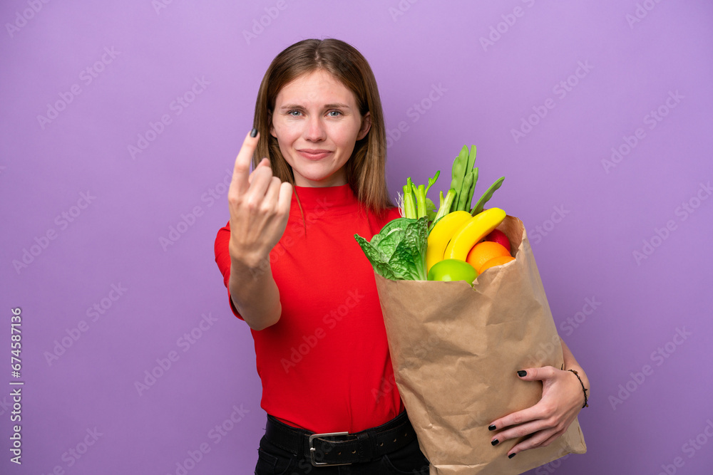 Young English woman holding a grocery shopping bag isolated on purple background doing coming gesture Young English woman holding a grocery shopping bag isolated on purple background doing coming gesture