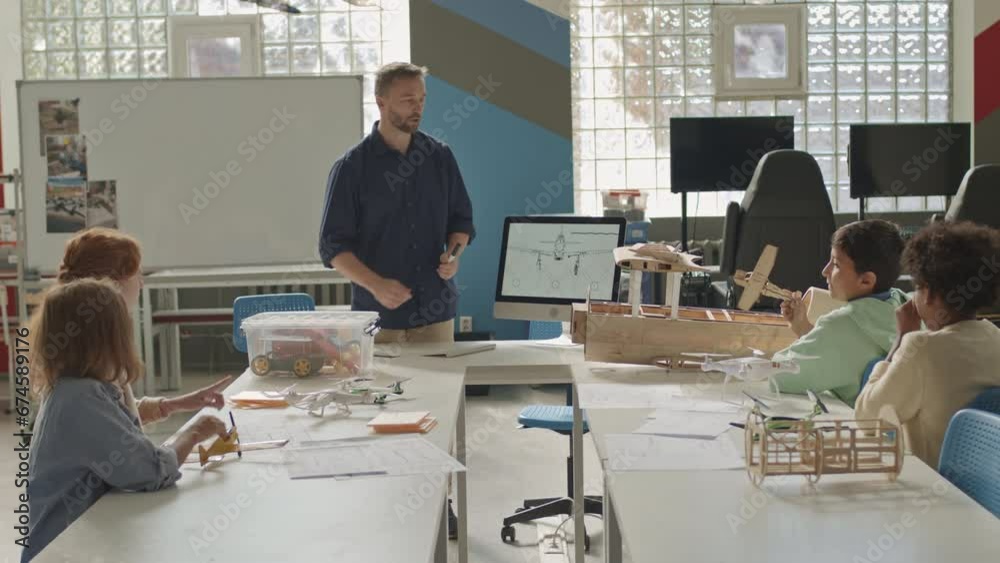 Medium shot of young Caucasian male teacher pointing at drawing scheme on desktop computer while explaining airplane construction to group of multiethnic students during engineering class