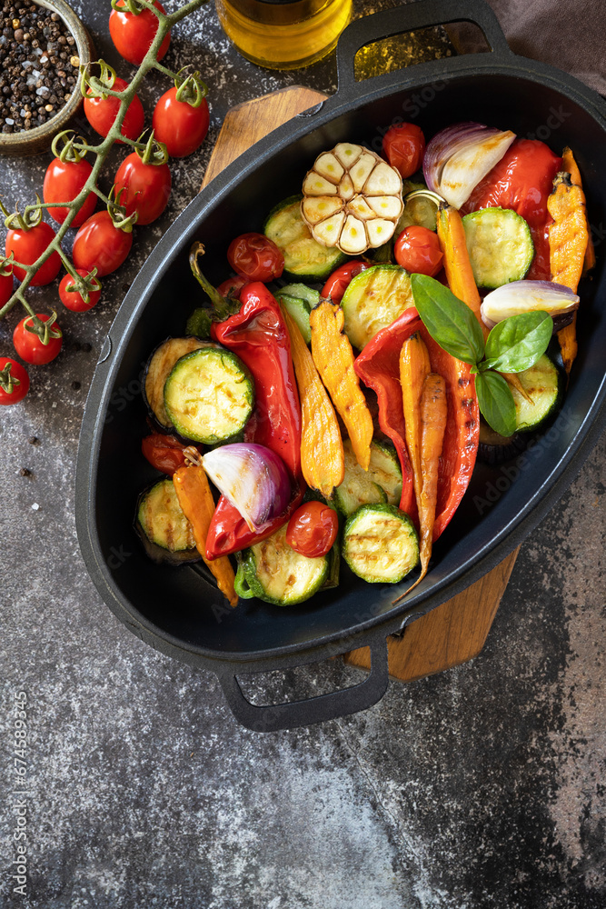 Grilled vegetables: red and yellow paprika, garlic, zucchini, eggplants, carrots in a frying pan on a dark stone background. View from above. Copy space. Grilled vegetables: red and yellow paprika, garlic, zucchini, eggplants, carrots in a frying pan on a dark stone background. View from above. Copy space.