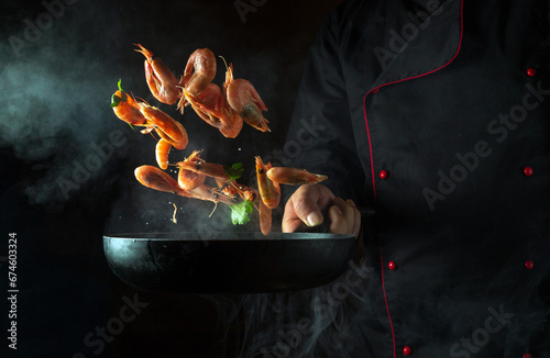 Fototapeta Naklejka Na Ścianę i Meble -  The chef cooks shrimp in a hot pan with steam. The concept of cooking seafood or healthy vegetarian food. Freeze in motion on a black background with free space for a recipe or menu