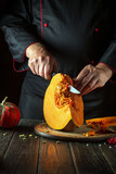A chef cuts an orange pumpkin into slices with a knife on a wooden cutting board before preparing porridge for breakfast. Autumn food concept in cozy dark kitchen