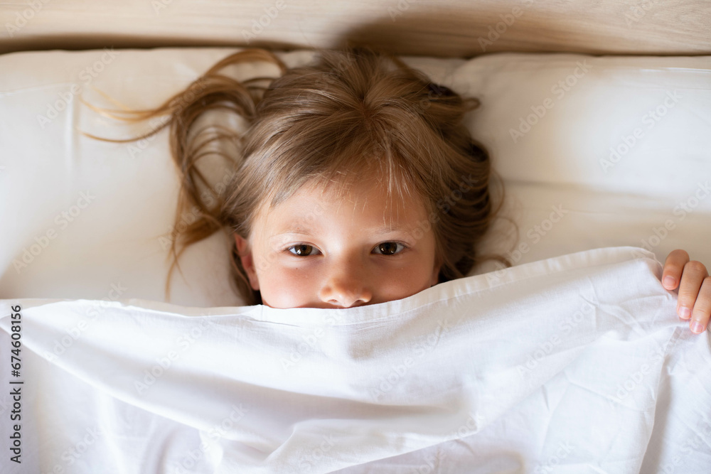 A charming happy little girl with long hair is lies on a white bed under a white blanket and laughs at the morning. It's perfect for projects related to family, childhood, or relaxation.Top view photo A charming happy little girl with long hair is lies on a white bed under a white blanket and laughs at the morning. It's perfect for projects related to family, childhood, or relaxation.Top view photo