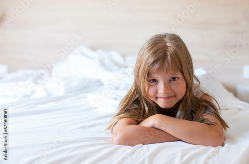 A little happy girl with long hair is lies on a white comfortable bed and laughing look front to the camera at the morning. It's perfect for projects related to family, childhood, or relaxation.