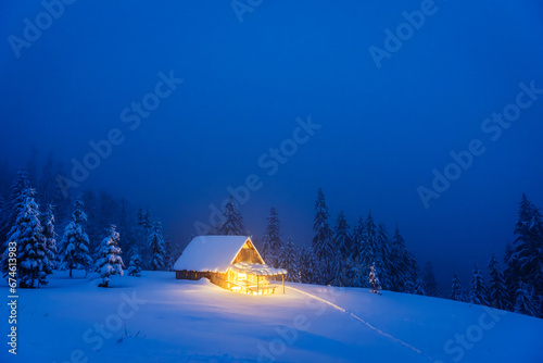 A winter landscape featuring an isolated wooden cabin and snow-covered fir trees on a mountain meadow deep within the forest. Christmas postcard. Snowy mountains forest