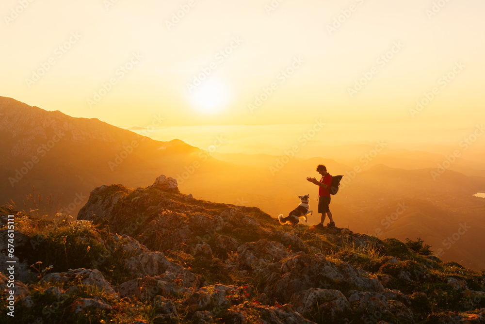 Mountaineer man with backpack plays with his border collie dog while hiking in the mountains at sunset. Mountain sports and adventure