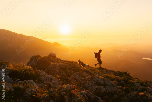 young man in silhouette descending a mountain peak at sunset with his dog. Sport, adventure and hiking in the mountains.