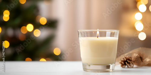A burning candle in a glass jar on white table with blurred Christmas lights in background