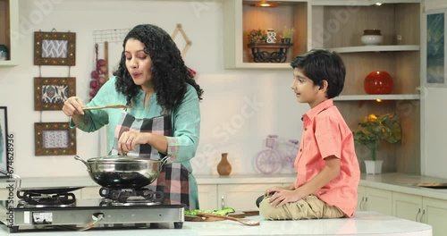 Indian mom cooking food and get food tested to her son sitting on the kitchen counter

