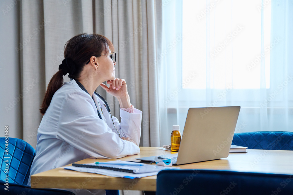 Confident serious mature woman doctor sitting at table with laptop computer Confident serious mature woman doctor sitting at table with laptop computer
