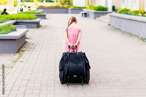Full length shot of a little girl walking and pulling a suitcase. Back view of a little girl in a pink dress traveling with a big bag. A child walks and rolls a suitcase on wheels along the sidewalk.