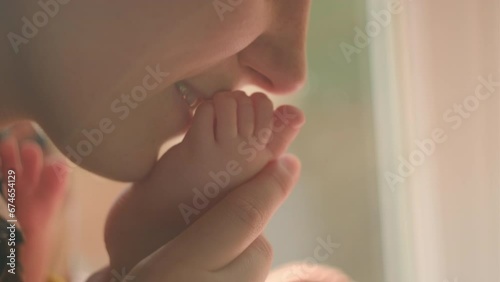Close up of cheerful young mom gently kiss little feet in warm sunlight sitting on sill at home, tiny toes of newborn baby child. Tender moments with kid, mother and offspring connection, care concept