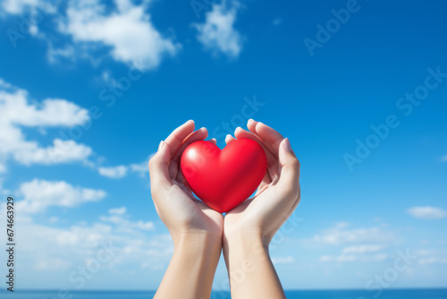 A woman s hand wrapped around a red heart is reaching towards the blue sky. A concept suitable for celebrating happiness and being grateful for happiness  as well as a heart of kindness and warmth.