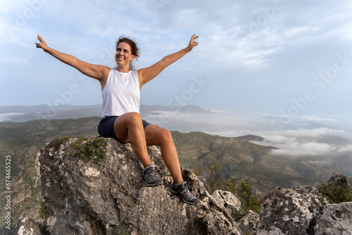A young brunette caucasian woman with her arms open in joy sitting on a rock on the top of a mountain