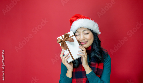 Pretty smiling asian woman in warm christmas sweater and santa hat holding gift box as present with red background for season celebration