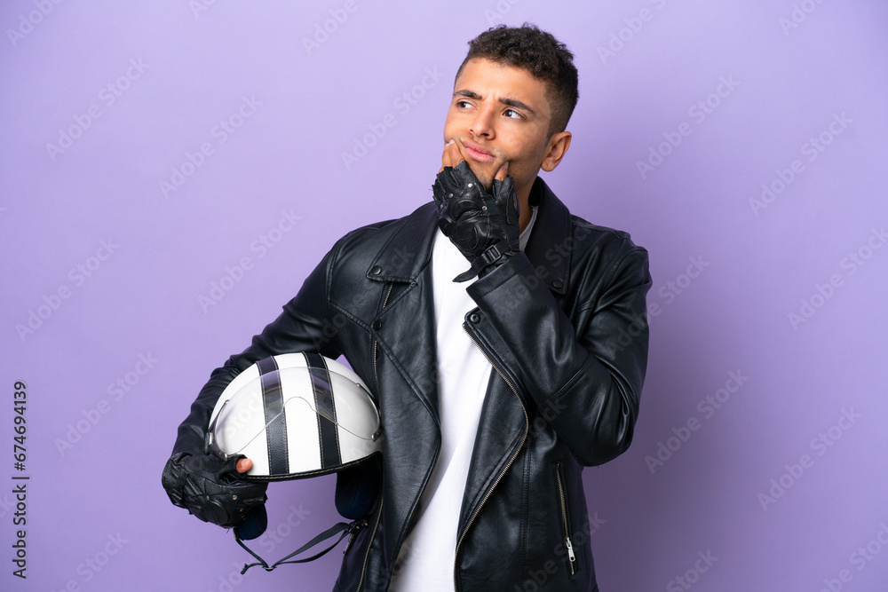 Fototapeta premium Young Brazilian man with a motorcycle helmet isolated on purple background having doubts