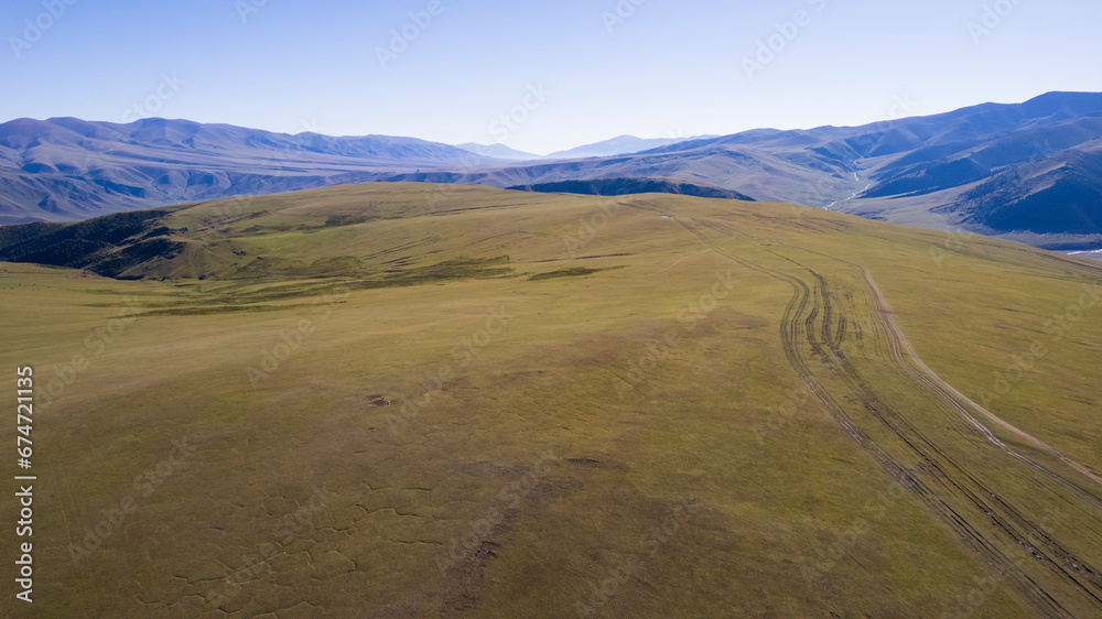 A white SUV is driving in the mountains through the fields. Aerial view from a drone of huge white clouds, snowy mountain peaks and green fields. Dirty off-road with puddles. Splashes from the wheels