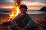 Happy young man sitting by the bonfire on the beach at sunset