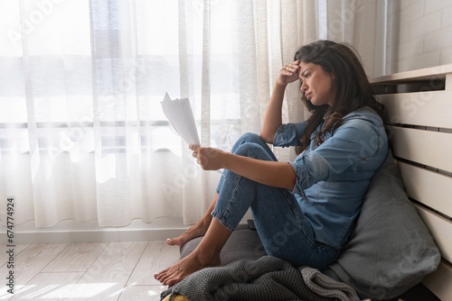 Stressed young woman holding financial papers sitting on the floor concerned about her finances photo
