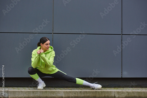young sportswoman in hoodie and leggings doing lunges near grey wall outdoors, energy and sport photo