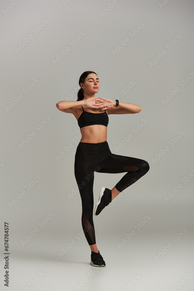 calm woman in active wear meditating and standing on one leg on grey background, beathing technique