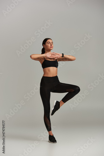 calm woman in active wear meditating and standing on one leg on grey background, beathing technique photo