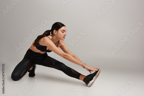 attractive woman in black active wear with fitness tracker on wrist stretching leg on grey backdrop