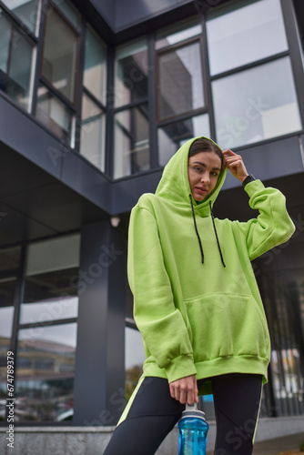 young sportswoman in oversized active wear wearing hood and standing with bottle with water outdoor © LIGHTFIELD STUDIOS
