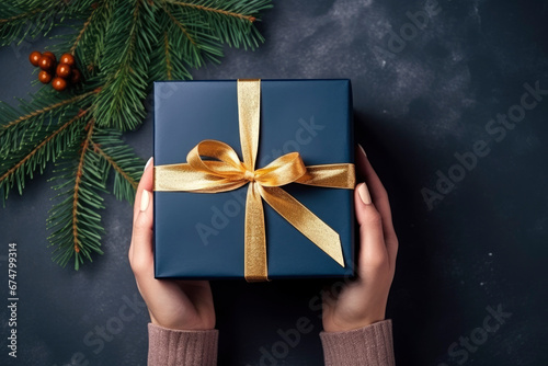 overhead view, womans hands holding a luxury gift box with gold bow against a dark blue background with christmas fir branches. Close up. New Year present.