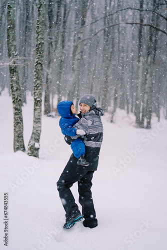 Smiling mother with a little girl in her arms walks through the snowdrifts in the forest under a snowfall