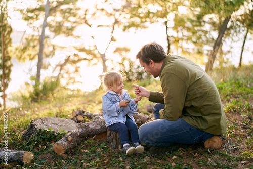 Dad holds out a blade of grass to a little smiling girl sitting on logs in the forest