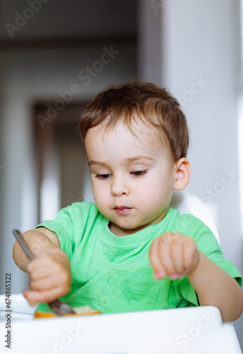 Curious Toddler Learning to Eat Independently. A little boy sitting in a high chair with a knife and fork in his hand